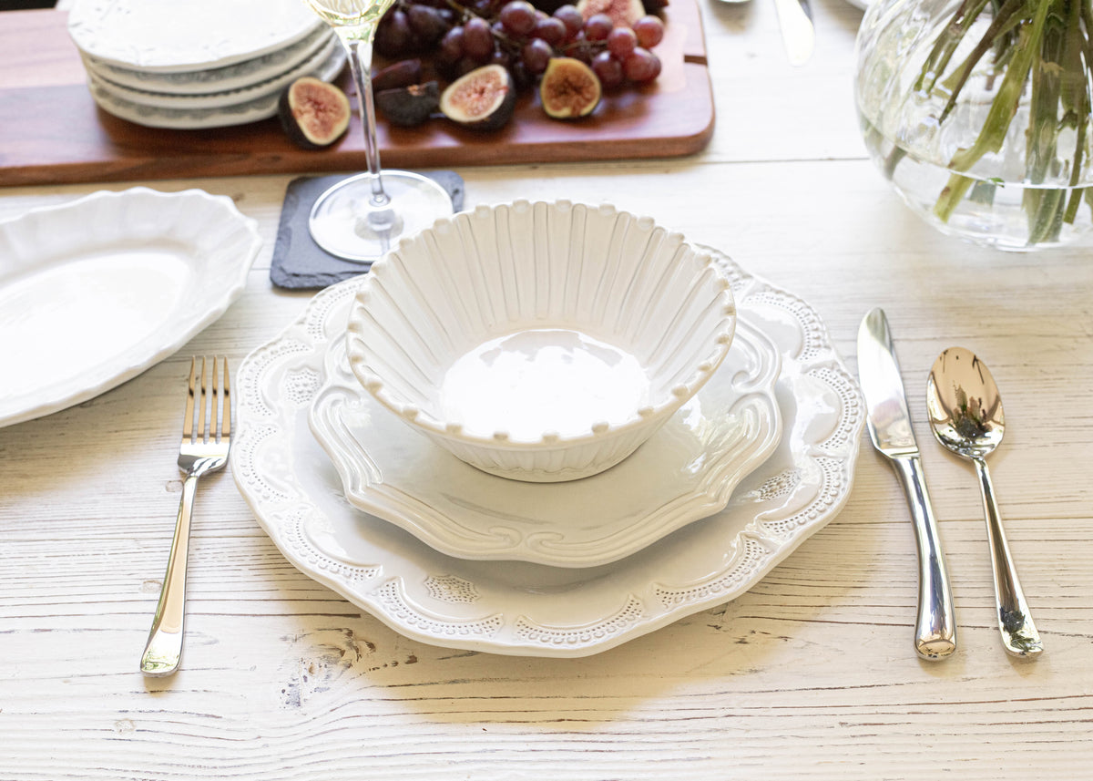 A white VIETRI Incanto dinner plate, salad plate, and cereal bowl all stacked on top of each other on a table with flatware on each side. 